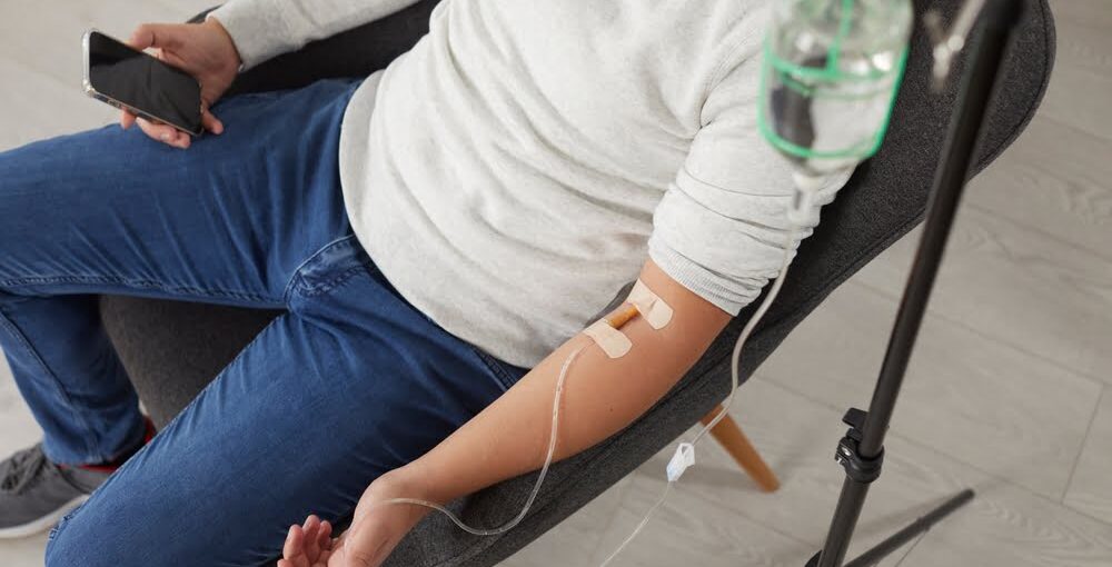 Person in casual clothing sitting in a chair receiving a magnesium IV therapy drip from a glass IV bottle stand