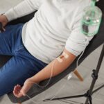 Person in casual clothing sitting in a chair receiving a magnesium IV therapy drip from a glass IV bottle stand