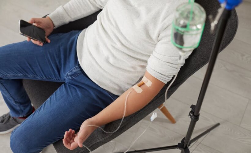 Person in casual clothing sitting in a chair receiving a magnesium IV therapy drip from a glass IV bottle stand