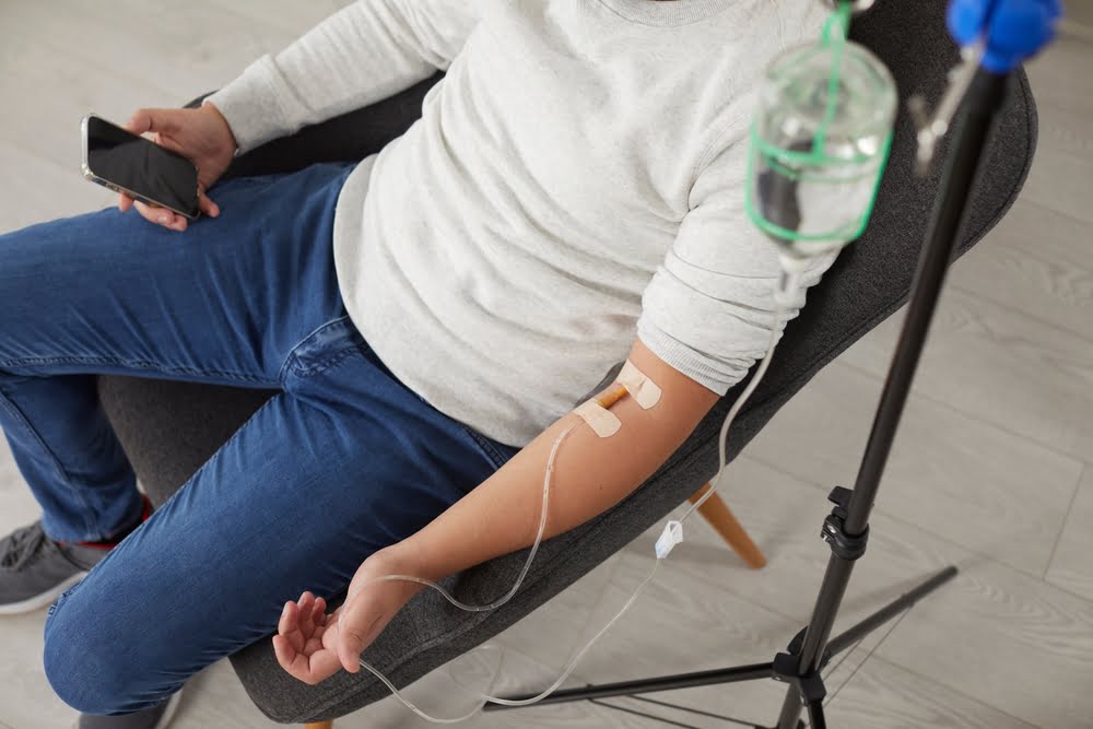 Person in casual clothing sitting in a chair receiving a magnesium IV therapy drip from a glass IV bottle stand