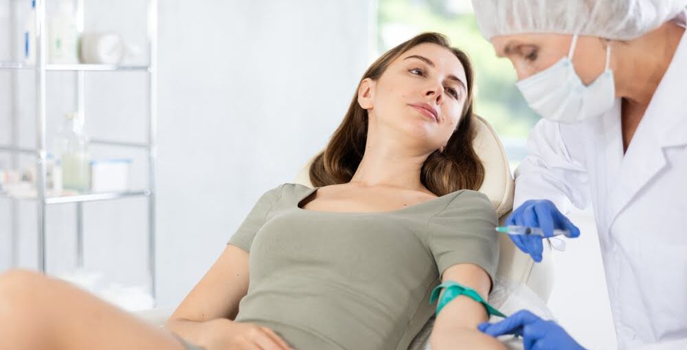 Young woman watching a masked practitioner prepare an IV line at her arm to deliver UVB therapy benefits through ultraviolet blood irradiation