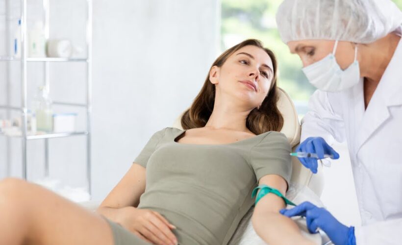 Young woman watching a masked practitioner prepare an IV line at her arm to deliver UVB therapy benefits through ultraviolet blood irradiation