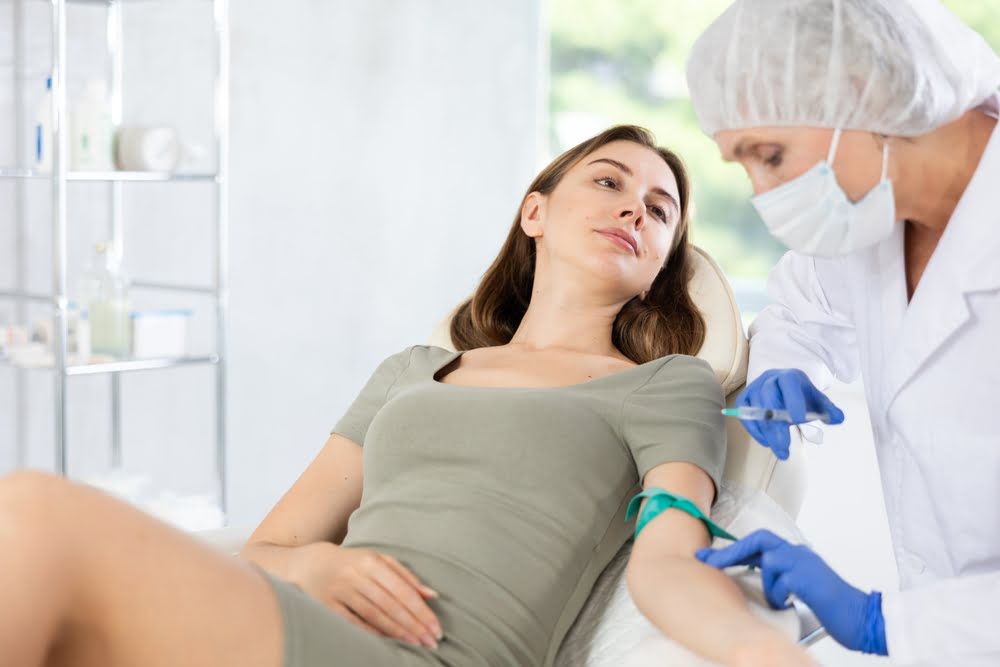 Young woman watching a masked practitioner prepare an IV line at her arm to deliver UVB therapy benefits through ultraviolet blood irradiation