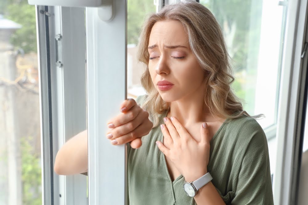 Woman standing by a window with hand on chest experiencing anxious distress, representing the biological and emotional burden that ozone therapy for anxiety aims to address