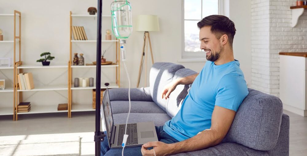 Smiling man in blue shirt relaxing on a couch with a laptop while receiving a vitamin C IV therapy drip in a bright home wellness setting