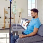 Smiling man in blue shirt relaxing on a couch with a laptop while receiving a vitamin C IV therapy drip in a bright home wellness setting