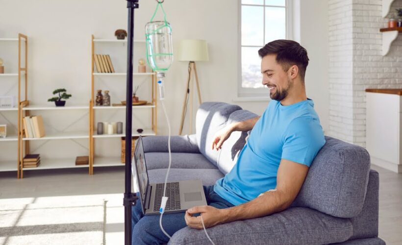 Smiling man in blue shirt relaxing on a couch with a laptop while receiving a vitamin C IV therapy drip in a bright home wellness setting