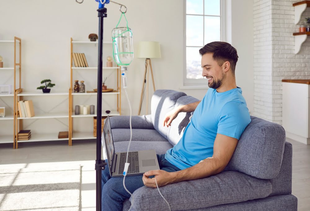 Smiling man in blue shirt relaxing on a couch with a laptop while receiving a vitamin C IV therapy drip in a bright home wellness setting